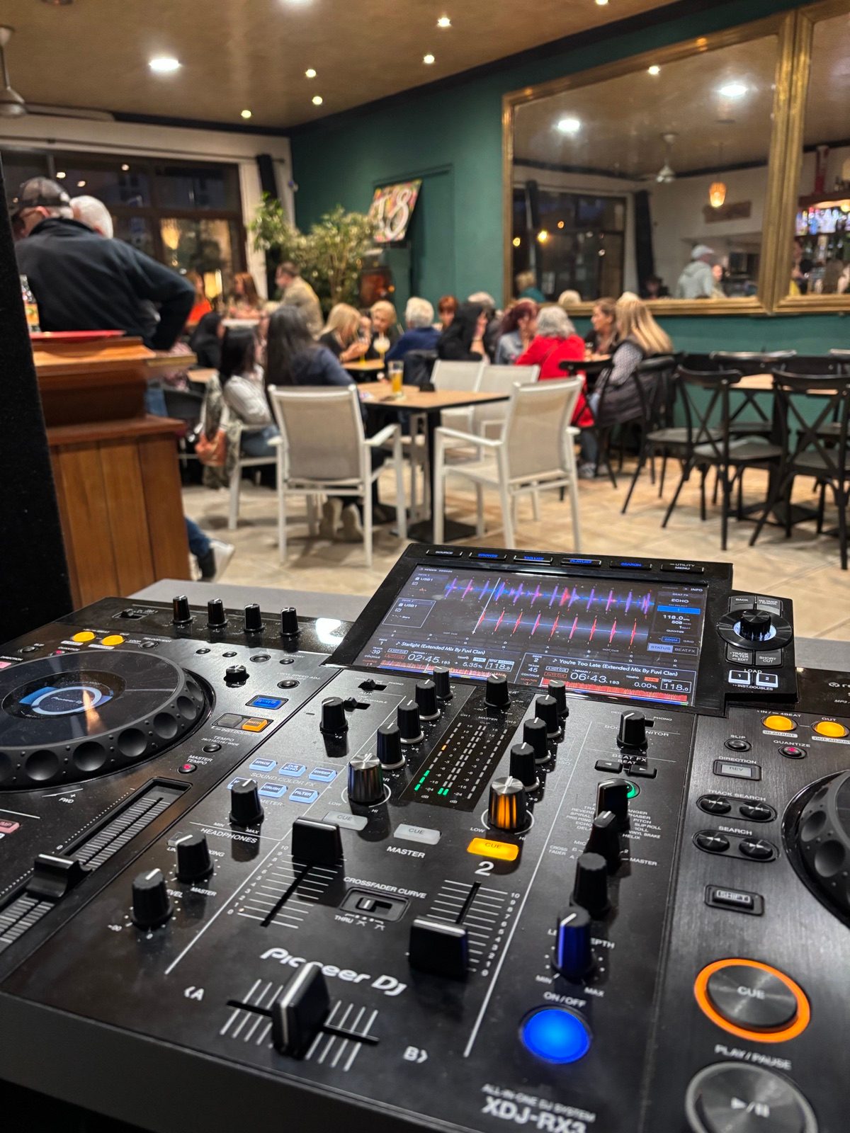Poste DJ au premier plan dans un bar, avec des clients attablés au fond de la salle
