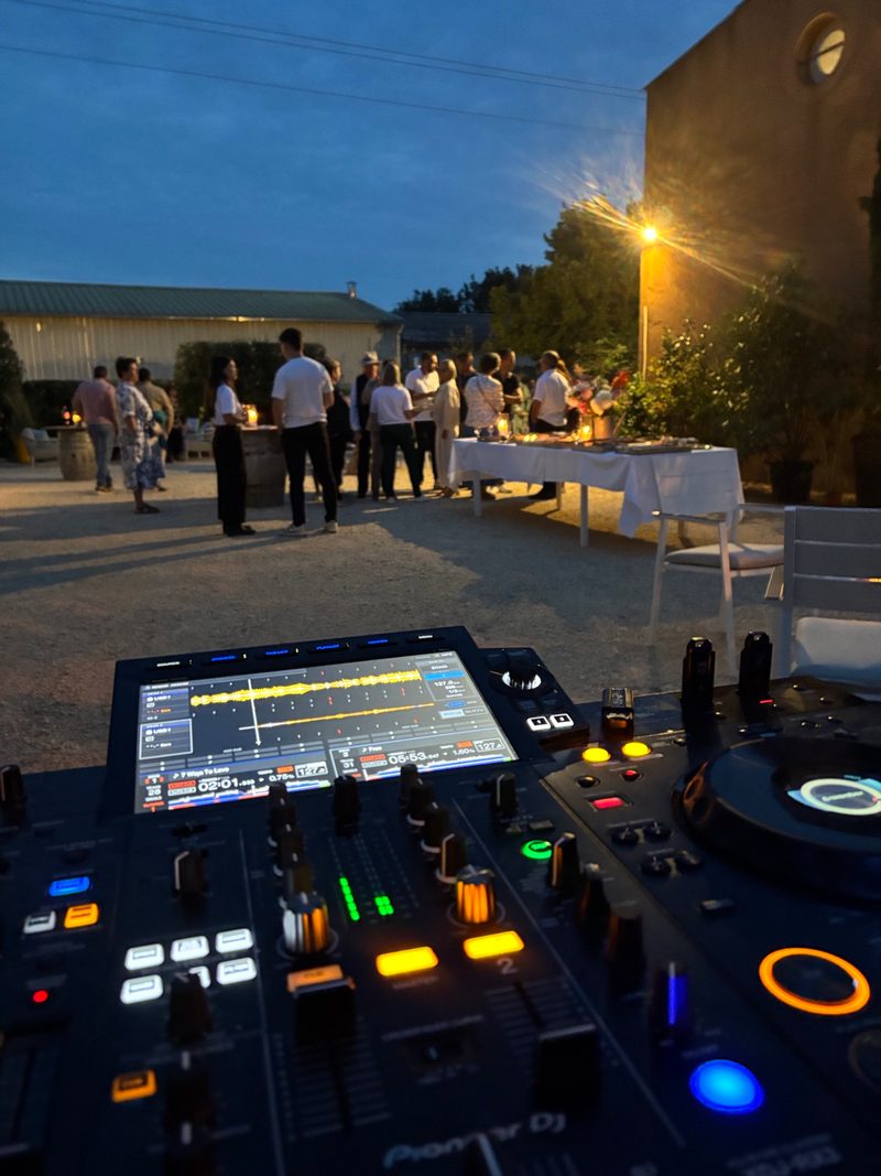Poste DJ allumé à la tombée de la nuit, invités debout autour d'un buffet en plein air