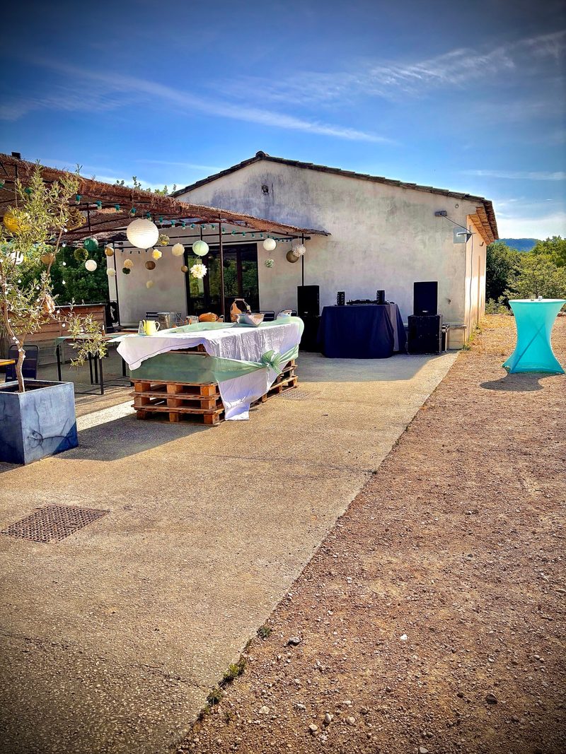 Terrasse d'une bâtisse en plein jour, avec tables nappées, lampions sous la pergola et poste DJ installé contre le mur